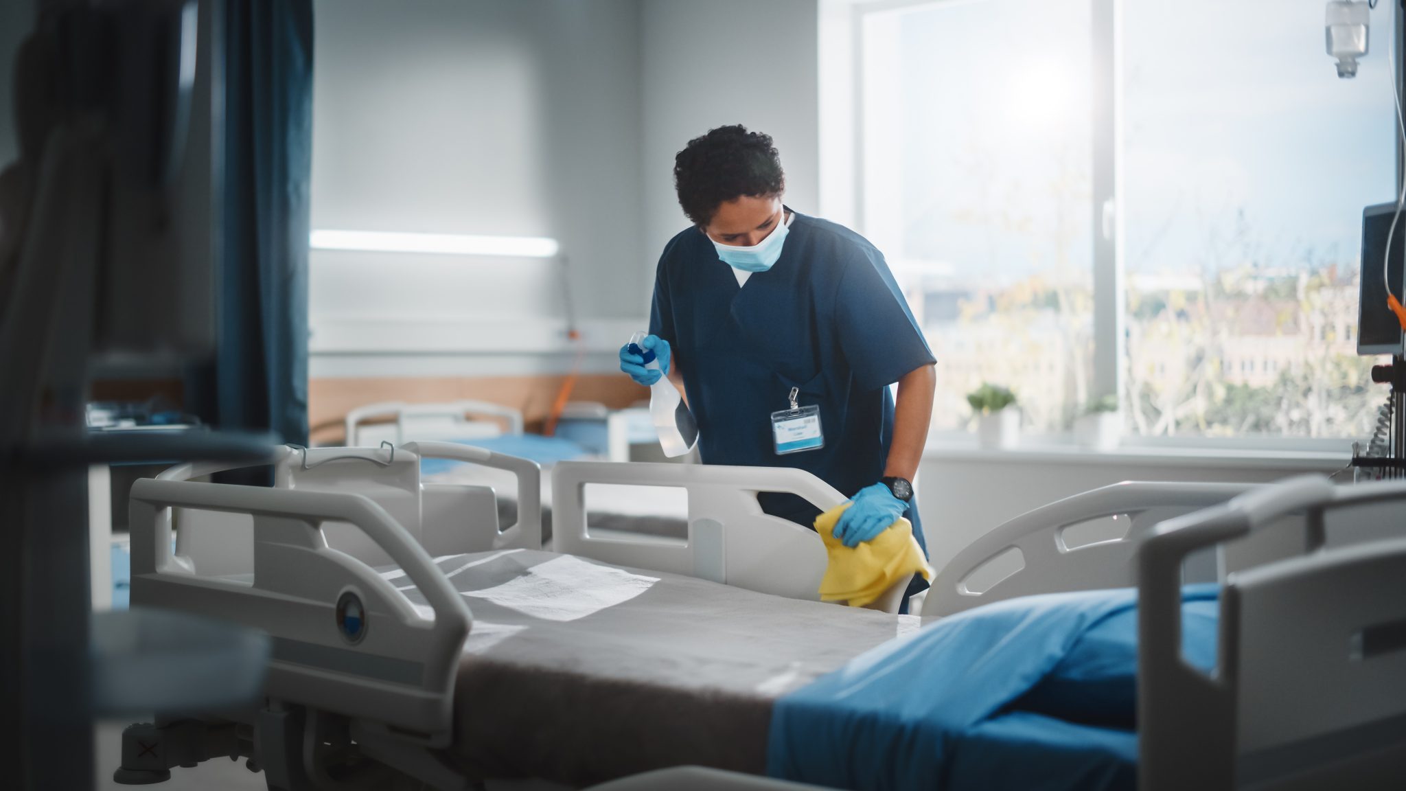 Healthcare worker in a mask and gloves cleaning a hospital bed rail